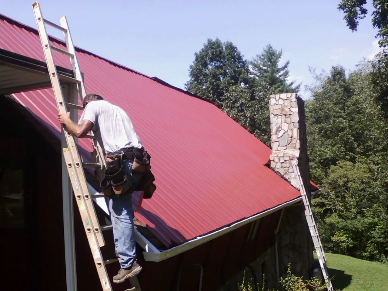 Roofer on a ladder beside a red metal roof and stone chimney — Soffit & Fascia Repair by Roofing Brothers in Woburn